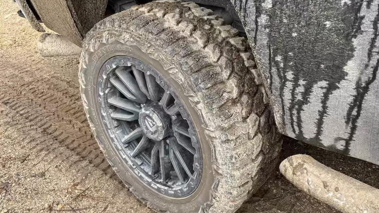 A muddied Deestone all-terrain tire mounted on a dark truck parked on a dirt surface.