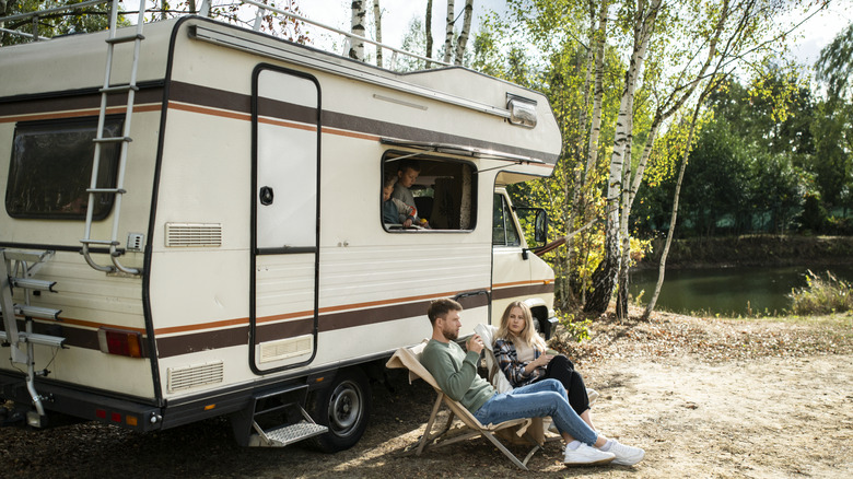 Two people sit in front of an RV while two children are inside it.