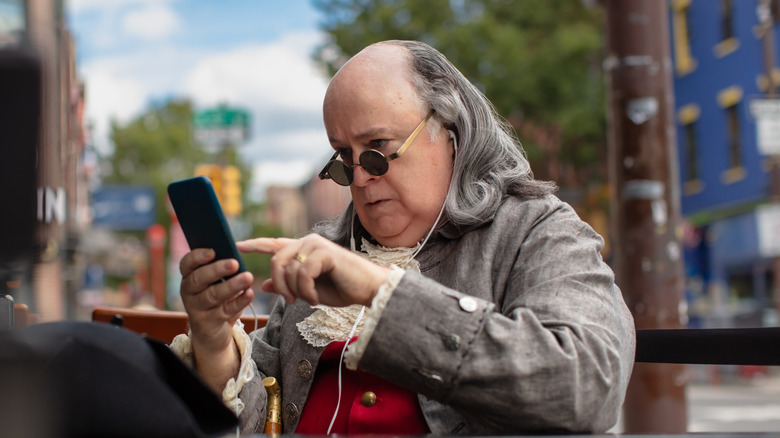 Benjamin Franklin impersonator sitting outside with a pair of headphones and a smartphone