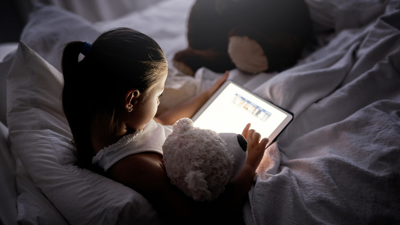 Child reading a tablet in bed next to a stuffed animal