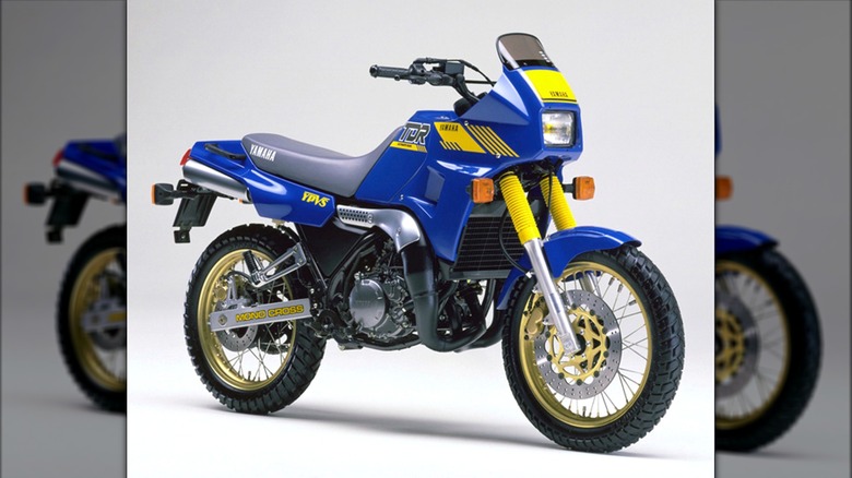 A 1998 classic blue and yellow Yamaha TDR250 motorcycle sits in a studio setting, showing off its rugged dual-sport frame, gold wheels, and tall stance.