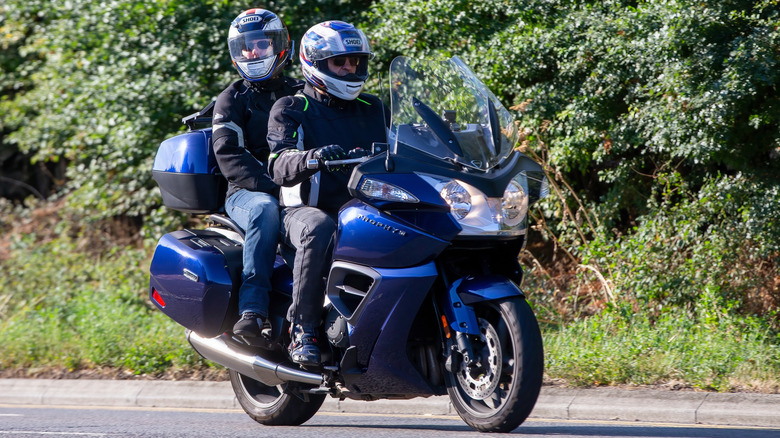 A biker and a passenger ride a Triumph Trophy 1200 SE on a sunny day