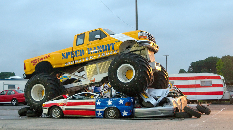 A yellow monster truck driving over various other vehicles