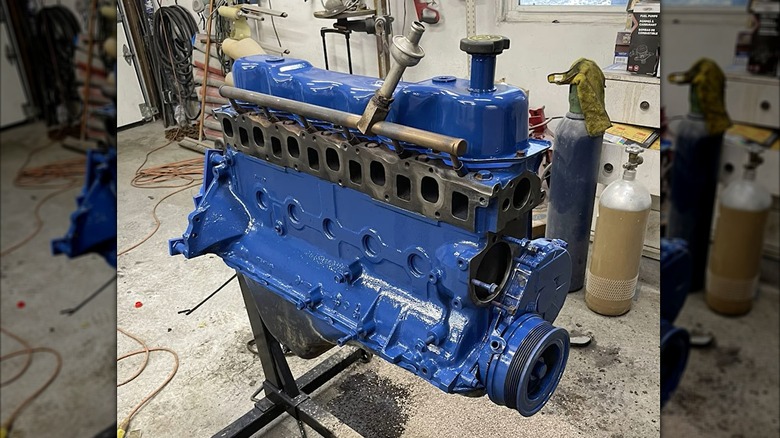 A freshly painted blue inline-six engine mounted on a stand in a workshop, with visible cylinder head ports and accessory mounting points.