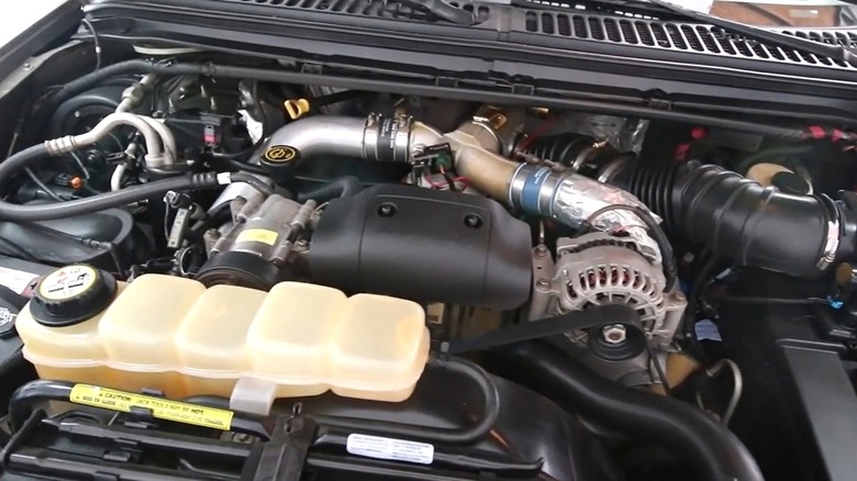 The engine bay of a pickup truck featuring a Ford 7.3L Power Stroke Diesel engine, with prominent turbocharger plumbing, alternator, and coolant reservoir visible under the open hood.