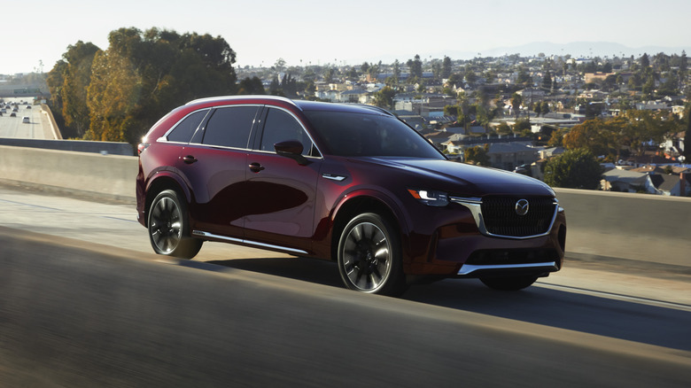 A maroon 2025 Mazda CX-90 PHEV driving through an urban environment, front 3/4 view