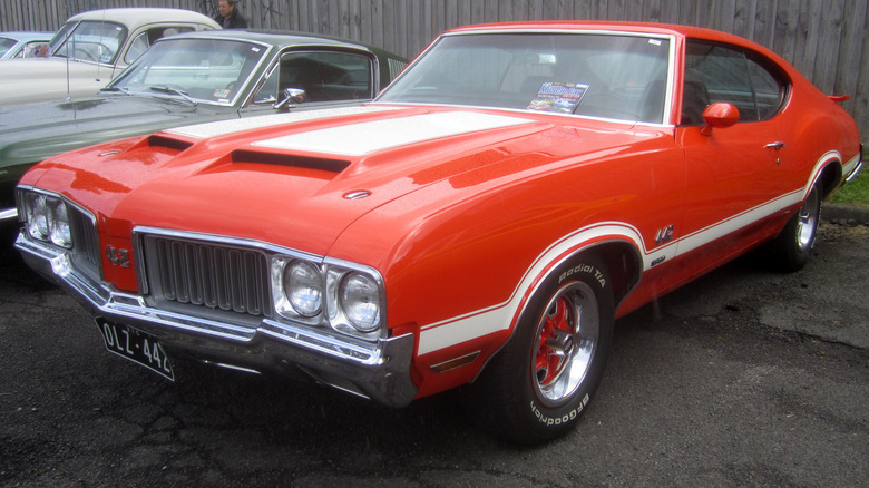 Red 1970 Oldsmobile 4-4-2 W30 coupe seen from front left parked with other vintage cars in front of a fence