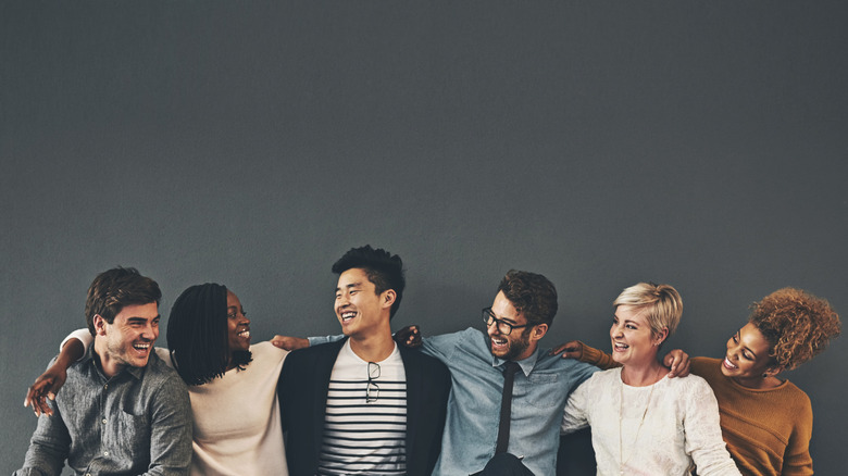 A group of people with arms on each other's shoulders and smiling, gray background.