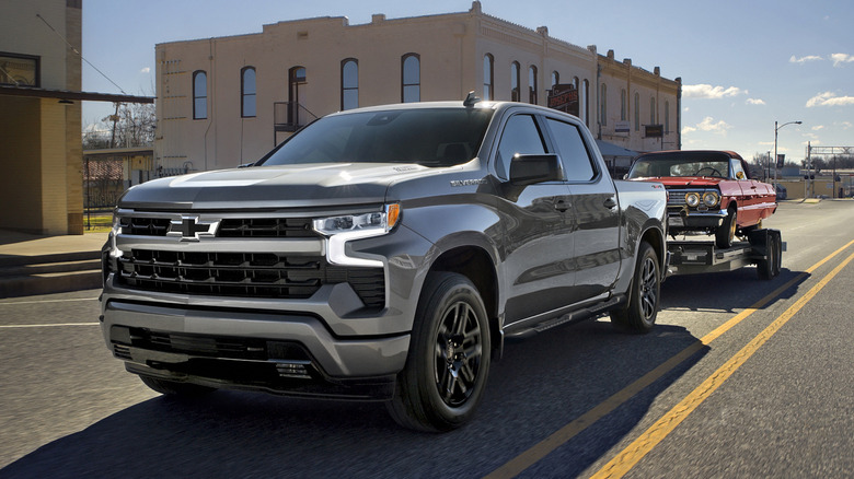 Silver Chevrolet Silverado 1500 towing a red muscle car on a trailer
