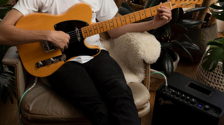 A person playing a guitar while sitting on a chair near an amp.