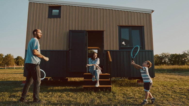 A man and a small boy pay tennis outside a small mobile home. A woman is sitting on the steps of their home smiling.