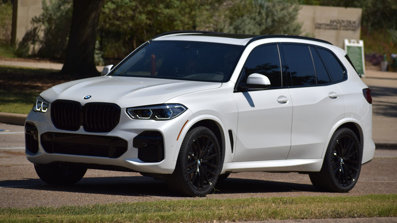 A white BMW X5 driving down suburban roads with green grass in front and a tree in the background.