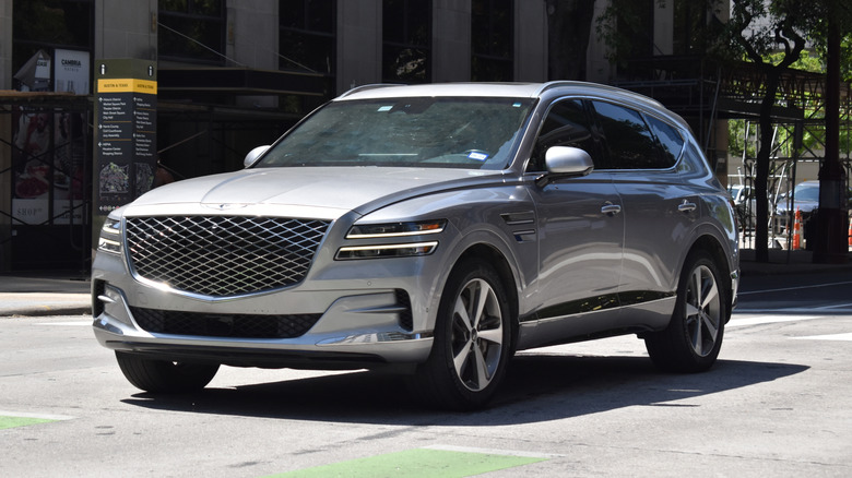 A silver Genesis GV80 cruising down an urban road with an industrial-looking building in the background.