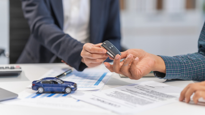 A shot of a salesman handing over car keys to a person sitting across from it, with car models and written forms on the table.