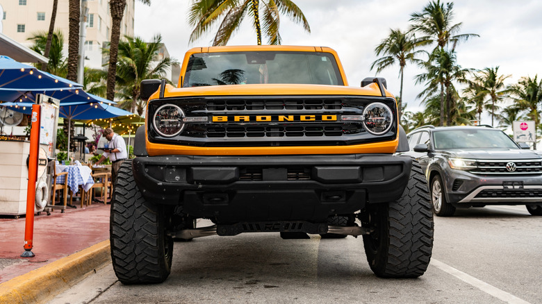 A front-facing shot of a modified yellow Ford Bronco SUV in an urban environment with palm trees in the background.
