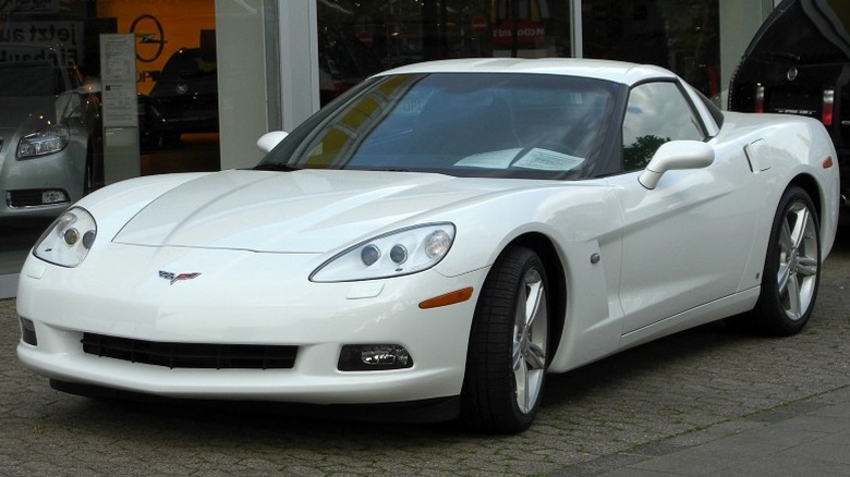 White C6 Chevrolet Corvette Coupe parked  on a paved surface outside a dealership building with other cars in the background