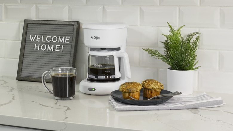 Mr. Coffee coffee maker on a counter with a welcome home sign, muffins, and a potted plant