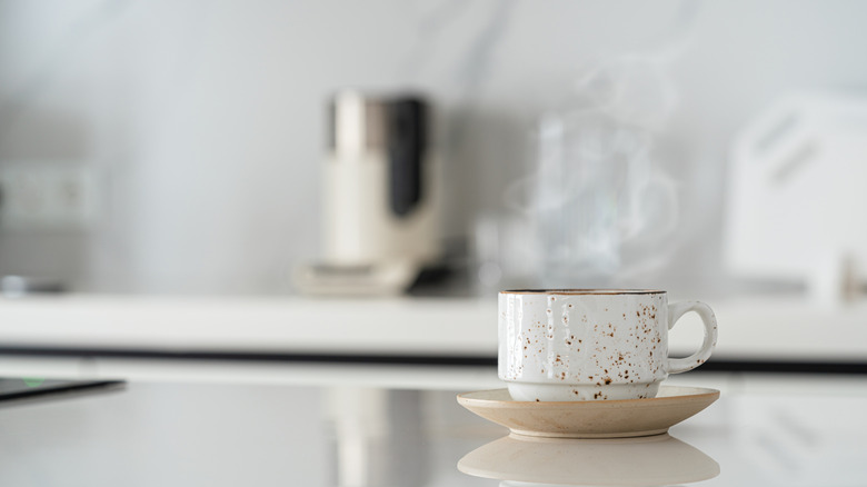 Coffee cup on a counter, coffee maker in the background, blurred.