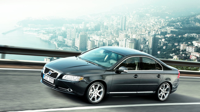 A black 2010 Volvo S80 executive sedan cruising along a highway with a city and river in the background.