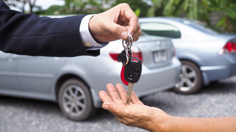 A person handing over car keys to another person with two silver cars on the back