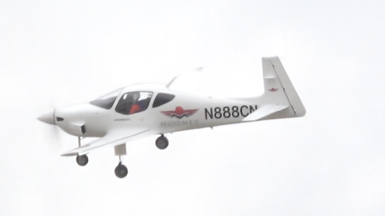 A blurry picture of a white Mooney M10 flying in the air, against a white background.