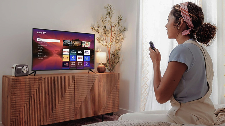 A woman sitting on a bed watching a Roku Plus Series TV.