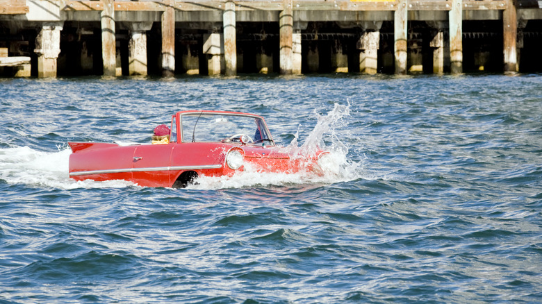 A red amphibious car floating through the water with a structure in the background.