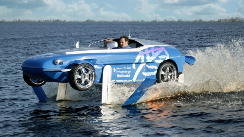 Rinspeed Splash hydrofoil amphibious car skimming across the water surface with two passengers, showcasing its water-lifting hydrofoil technology.