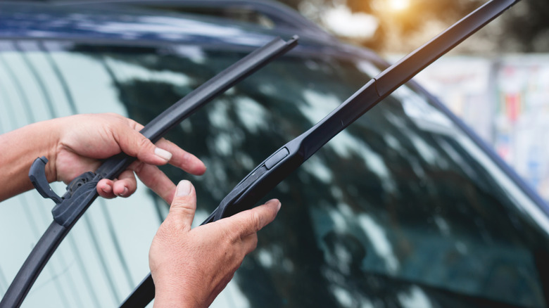 A pair of hands hold a pair of replacement wiper blades with a car windshield in the background.