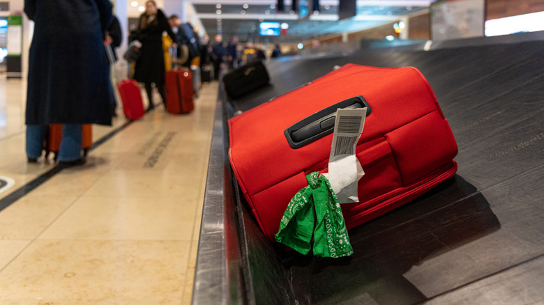 A red suitcase with a green and white piece of cloth tied around the handle, sitting on an airport terminal conveyor belt.
