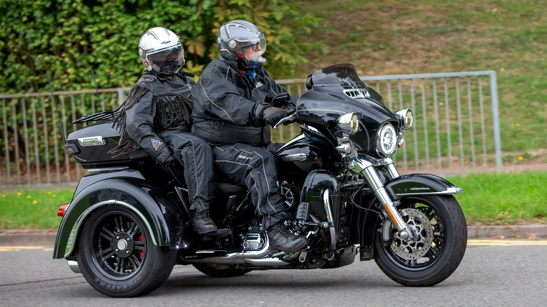 Two bikers on a Harley-Davidson Trike traveling on a British road with bushes in the background