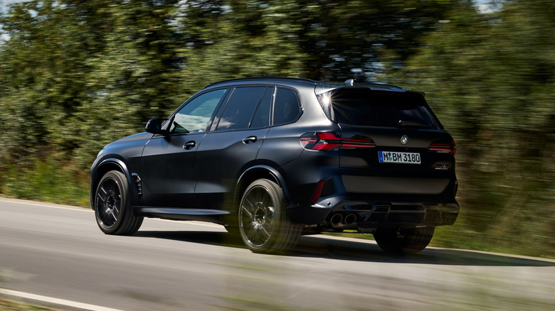 A rear-facing shot of a dark-colored BMW X5M driving with trees in the background.