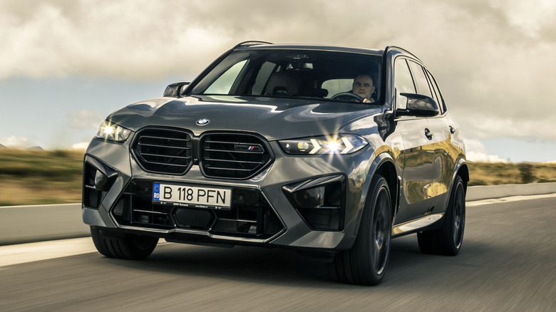 A front-facing shot of a dark-colored BMW X5M driving on open roads with headlights turned on.