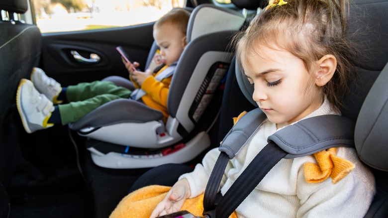 Cute siblings sitting in baby booster car seats and using mobile phones during the road trip