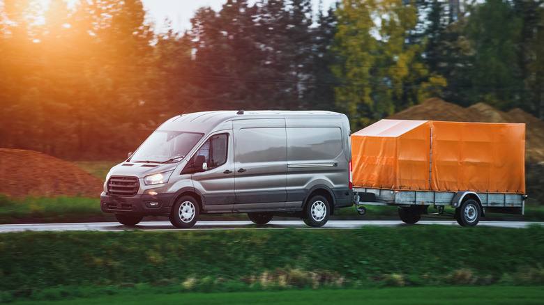 A Ford Transit pulling a covered trailer through a forest road, front 3/4 view