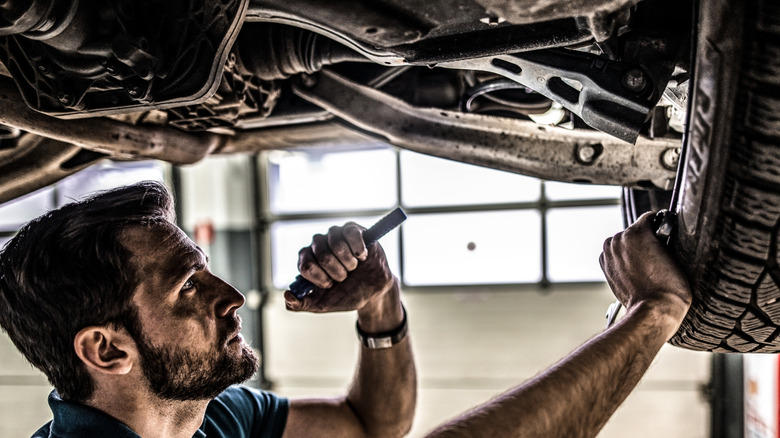A mechanic uses a flashlight to inspect the suspension on the underside of a car.