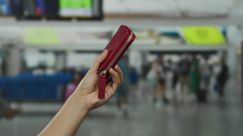 A hand holding a red wallet with a blurry airport terminal in the background