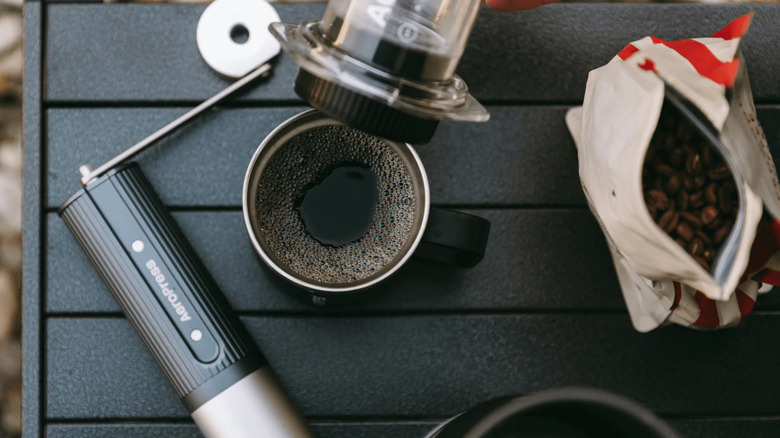 Black coffee in a metal mug with an Aeropress machine and Aeropress manual grinder on a table with coffee beans.