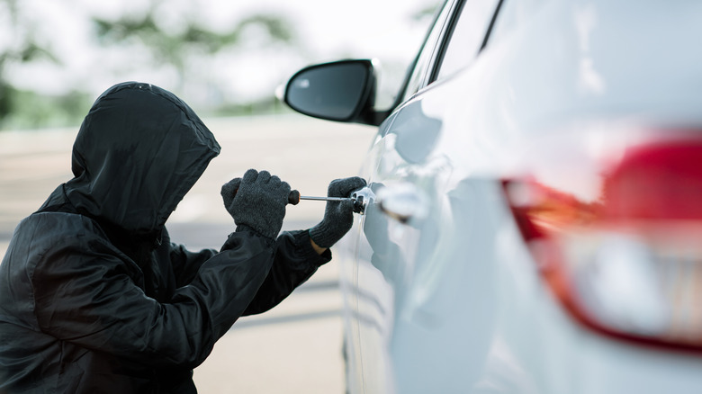 A man dressed in black with his face hidden attempting to break into a car with a screwdriver