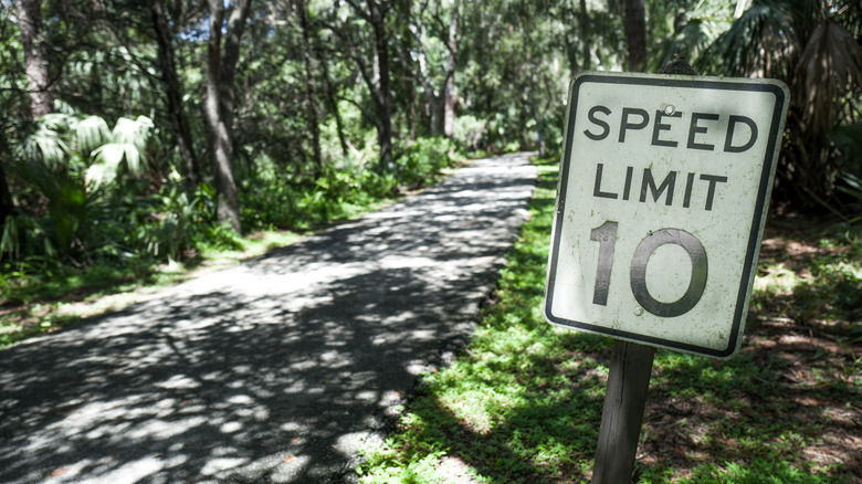 A 10 MPH speed limit sign on the side of a paved trail in a wooded area