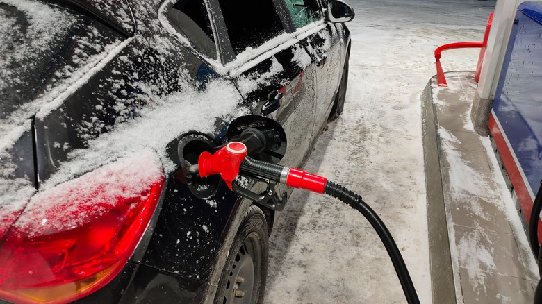 A snow-covered black car is filling up at a gas station on a winter night