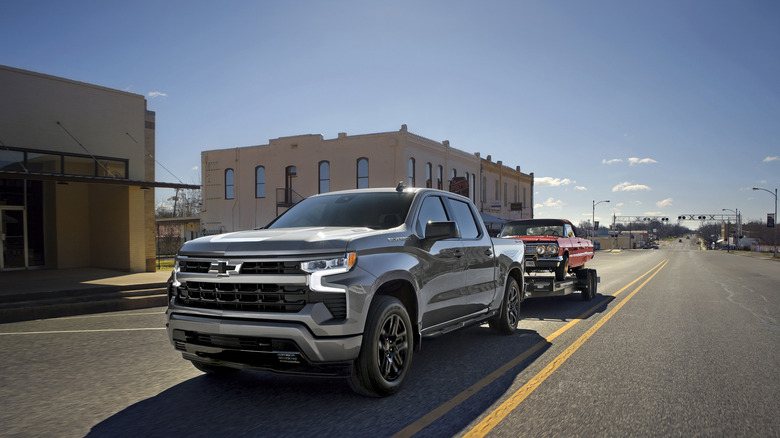 Silver 2026 Chevrolet Silverado towing red 1963 Chevrolet Impala on trailer on two-lane road