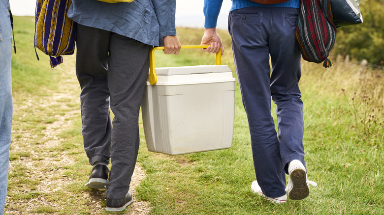 Two adults carrying a light-colored cooler between them on a grassy path
