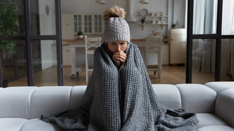 A cold woman at home on a sofa wrapped in a gray blanket and wearing a gray winter hat