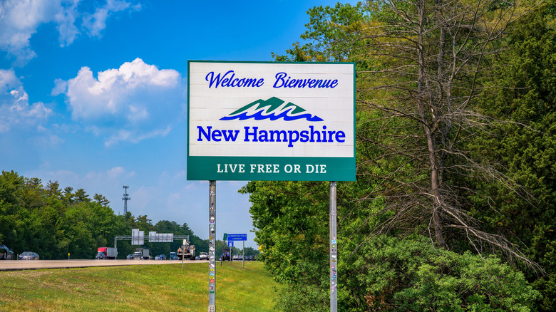 A white road sign reading "Welcome to New Hampshire," with greenery next to the road in the background.