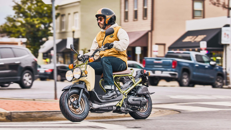 A rider in a helmet and yellow jacket riding a yellow Honda Ruckus through an intersection