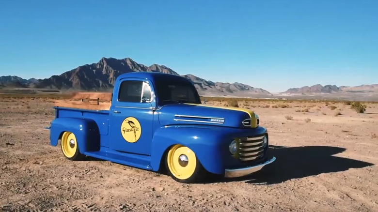A bright blue vintage pickup truck with yellow wheels and a Glasurit logo on the door parked in the desert.