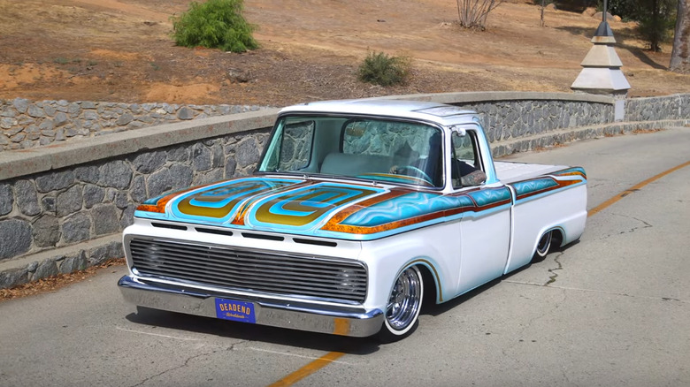 A custom white lowrider pickup truck with turquoise, gold, and brown patterns on the hood and sides, parked on a narrow stone-lined road.