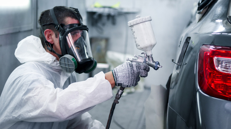An auto painter wearing a full-face respirator, protective suit, and gloves sprays paint onto the side of a gray car.