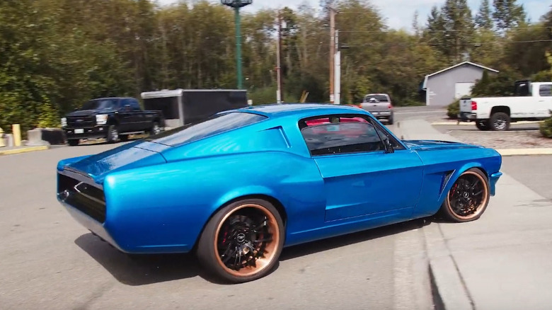 A bright blue classic Ford Mustang fastback with bronze wheels pulls into a parking lot.
